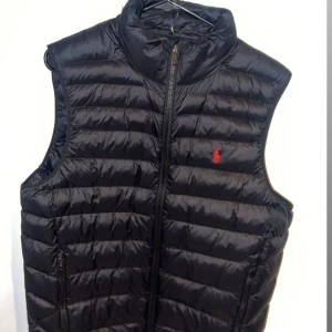 Svart dunväst från Polo Ralph Lauren - Säljer en svart dunväst från Polo Ralph Lauren med horisontella quiltade paneler och dragkedja framtill. Västen har två fickor med dragkedja och klassisk röd logga broderad på bröstet. Perfekt att slänga över en hoodie för en clean look.