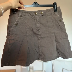 Brun kortkjol från H&M L.O.G.G. - Snygg brun kortkjol från H&M L.O.G.G. med coola fickor både fram och bak samt dekorativa knappar. Kjolen har bälteshällor och är tillverkad i bomull, vilket gör den skön och lätt att bära. Perfekt för dig som gillar en avslappnad och trendig stil. Den skiftar färg till lite mer militärgrön i dagsljus.