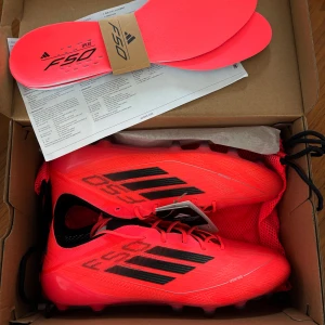 Adidas F50 Speed röda fotbollsskor - Snygga Adidas F50 Speed fotbollsskor i en riktigt skarp röd färg med svarta detaljer och logga. Skorna har snörning, platt sula med dobbar och är tillverkade i lätt syntetmaterial för maxad fart på planen. Perfekta för dig som vill sticka ut på matchen.