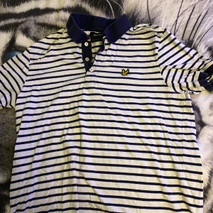 Randig pikétröja från Lyle & Scott - Snygg randig pikétröja från Lyle & Scott i vitt och marinblått. Klassisk krage med knappar och broderad gul logga på bröstet. Perfekt för en stilren och avslappnad look. Tillverkad i mjuk bomull och har korta ärmar.