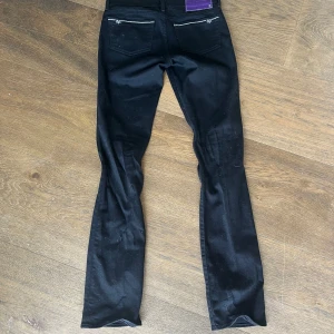Svarta jeans från Denim by Victoria Beckham - Svarta jeans med raka ben från Denim by Victoria Beckham. Snygga silverdetaljer vid bakfickorna och lila patch i midjan. Tillverkade i stretchig denim av 99% bomull och 1% spandex för en bekväm passform.