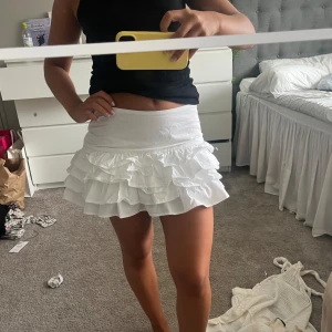 Vit volangkjol med flera lager - Supersöt vit kortkjol med flera lager volanger som ger en fluffig och lekfull look. Kjolen har bred midja och är perfekt för dig som vill sticka ut med en feminin och trendig stil. Lätt och luftig, passar perfekt till varma dagar.