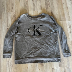 Grå sweatshirt från Calvin Klein Jeans. - Snygg grå sweatshirt från Calvin Klein Jeans med stort svart CK-tryck på bröstet. Tröjan har rund hals, långa ärmar och är tillverkad i mjuk bomullsmix. Perfekt för en chill och stilren look.