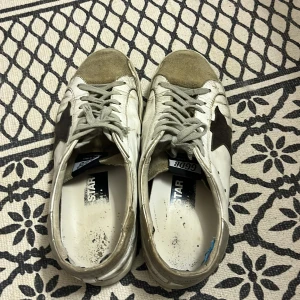 Golden Goose vita sneakers med stjärna - Snygga vita sneakers från Golden Goose med grå mockadetaljer på tån och grå snörning. Skorna har en svart stjärna på sidan och klassisk låg profil. Ovandelen är i skinn och mocka, vilket ger en cool kontrast. Perfekta för dig som gillar streetstyle.