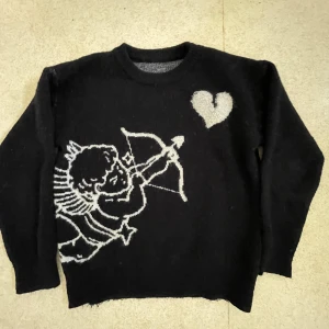 Svart stickad tröja med ängel och hjärta - Svart stickad tröja med vit illustration av en ängel med pilbåge och ett hjärta på bröstet. Tröjan har rund halsringning och långa ärmar. Perfekt för dig som gillar unika och lite edgy motiv.