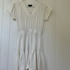 Vit kortklänning från Alyssa Thomas - Supersöt vit kortklänning från Alyssa Thomas med v-ringning, korta puffärmar och markerad midja. Klänningen har volangdetaljer i kjolen och är gjord i mjuk bomull, perfekt för varma dagar. Enkel och stilren design som känns fräsch och trendig. Köpt i en butik utan för New York