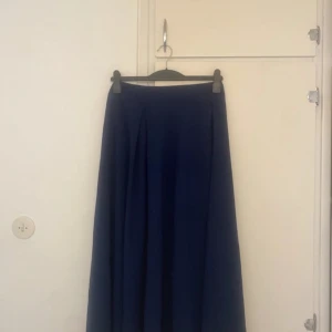 Mörkblå långkjol med veck - En stilren mörkblå långkjol med snygga veck framtill. Kjolen har en klassisk, vid siluett och hög midja som ger en tidlös look. Perfekt att matcha med en enkel topp eller stickad tröja för en chill och snygg outfit.