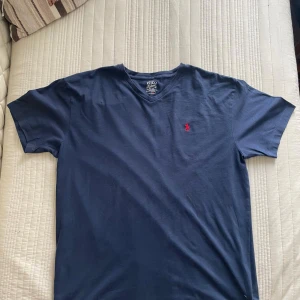 Blå t-shirt från Polo Ralph Lauren - Snygg marinblå t-shirt från Polo Ralph Lauren med klassisk röd broderad logga på bröstet. T-shirten har v-ringning och är tillverkad i mjuk bomull. Perfekt basic-plagg med normal passform och korta ärmar.