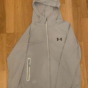 Ljusgrå vindjacka från Under Armour - Snygg ljusgrå vindjacka från Under Armour med huva och reflekterande detaljer. Jackan har dragkedja framtill, en praktisk ficka med dragkedja på sidan och logga på bröstet. Perfekt för dig som gillar sportig stil och vill sticka ut med coola detaljer.