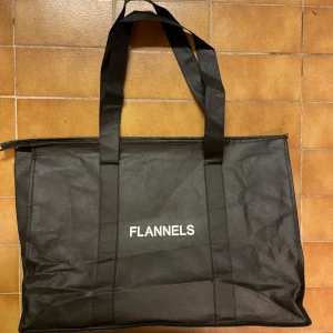 Stor svart shoppingväska från Flannels - En stor svart shoppingväska från Flannels med dragkedja upptill och långa handtag. Väskan är tillverkad i syntetmaterial och har enkel, stilren design med vit logga framtill. Perfekt när du behöver extra plats för grejerna.