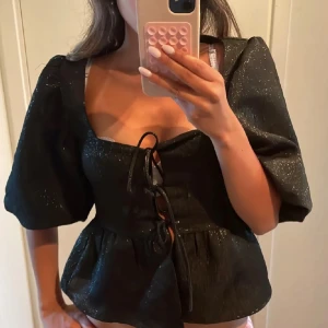 Svart glittrig blus med snörning - Supersnygg svart blus med glittrigt tyg och puffiga ärmar. Toppen har en djup urringning och snörning framtill som ger en cool och trendig look. Perfekt för dig som vill sticka ut lite extra.
