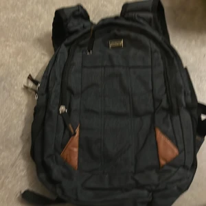Svart ryggsäck från Canyon - Snygg svart ryggsäck från Canyon med flera fack och dragkedjor. Väskan har bruna detaljer i hörnen och justerbara axelremmar. Perfekt för skola eller vardag, med slitstarkt tyg och praktisk design.