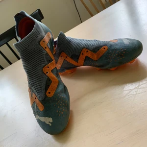 Puma Future Ultimate FG/AG Neymar JR Pro fotbollsskor grå och orange - Säljer ett par gråa och orangea fotbollsskor från Puma med stickad strumpliknande krage och coola detaljer i orange. Skorna har mönstrad ovandel och klassisk Puma-logga framtill. Perfekta för dig som vill sticka ut på planen. Väger runt 230 g per sko. Har andvänts i runt omkring ett halvår men är fortfarande i mycket bra skick. Priset är absolut inte hugget i sten och får gärna ändras om så önskas kontakta mig bara för fler bilder om så är fallet också.