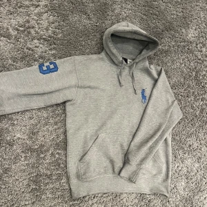 Grå hoodie från Ralph Lauren - Säljer en stilren grå hoodie från Ralph Lauren med blå logga på bröstet och stort nummer 3 på ärmen och ryggen. Tröjan är bra skick förutom att den är lite sliten på smörerna längst ut annars i bra skick!