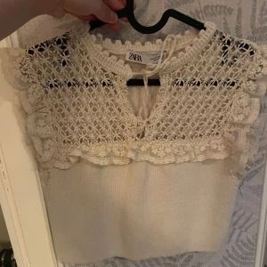 Vit spetsblus från Zara - Superfin vit blus från Zara med virkade spetsdetaljer och volanger vid axlarna. Toppen har knytning vid halsen och är ärmlös. Perfekt för dig som gillar romantisk och bohemisk stil.