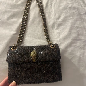 Svart paljettväska från Kurt Geiger - Snygg svart axelväska från Kurt Geiger med glittriga paljetter och kedjerem i antikguld. Väskan har ett dekorativt spänne framtill och en metallbricka med logga på baksidan. Perfekt för dig som vill sticka ut med en cool accessoar.