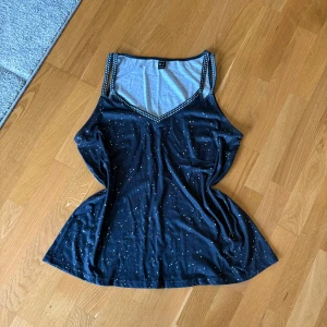 Svart linnetopp med prickiga band Sheilay - Snygg svart linnetopp från Sheilay med vita prickar på axelbanden och v-ringad hals. Toppen har ett stjärnliknande mönster över hela tyget och är tillverkad i mjuk bomullsmix. Perfekt för dig som gillar unika detaljer och bekväm passform.