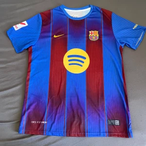 Barcelona tröja 25/26 Lamine Yamal - Säljer en officiell FC Barcelona matchtröja från Nike med Lamine Yamal och nummer 19 på ryggen. Tröjan har klassiska blå och vinröda ränder, gult Spotify-tryck på bröstet och UNHCR-logga bak. Kortärmad modell i lätt och ventilerande Dri-FIT-material.