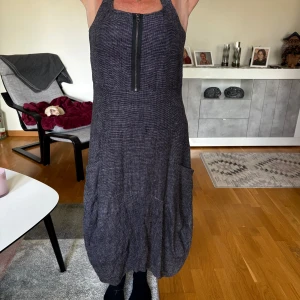 Mörkgrå långklänning med fickor - Säljer en mörkgrå långklänning med breda axelband och cool ballongformad nederdel. Klänningen har stora fickor på sidorna och en synlig dragkedja framtill som ger en edgy vibe. Materialet känns som bomull eller linne och tyget har en diskret struktur.