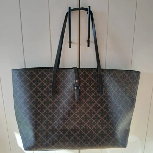 By Malene Birger - Stor shoppingväska från By Malene Birger med klassiskt grafiskt mönster i svart och brunt. Väskan har svarta långa handtag och ett spänne framtill. Utsidan är i syntetmaterial och insidan är beige. Perfekt för dig som vill ha plats med allt. Använd,  men är i mycket bra skick 