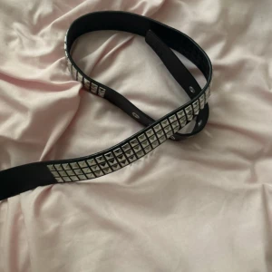 Svart nitarbälte med silverdetaljer - Säljer ett svart bälte i skinn med fyrkantiga silverfärgade nitar längs hela bandet. Bältet har en klassisk silverfärgad spänne och flera metallhål för justering. Perfekt accessoar för att ge din outfit en edgy vibe.
