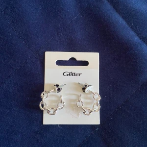 Silverfärgade örhängen från Glitter - Säljer ett par coola örhängen från Glitter i silverfärg. De är formade som små hoops med kedjelänkar, vilket ger en trendig och edgy vibe. Perfekta för att lyfta din stil med en modern touch.