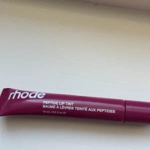 Rhode läppglans - Rhode Peptide Lip Tint i färgen Raspberry Jelly, en bärig och glossig läpprodukt i tub. Ger en hint av hallonrosa färg och återfuktar läpparna med peptider. Perfekt för dig som vill ha både färg och vård i ett. Tuben rymmer 10 ml och är smidig att ta med.