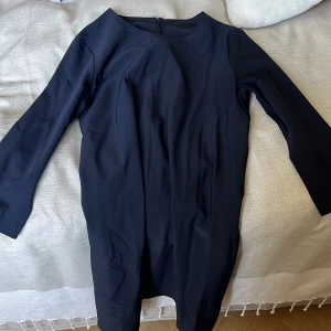 Mörkblå klänning från COS, stl 44 - En stilren mörkblå klänning från COS i storlek 44. Klänningen har lång ärm, rund halsringning och en rak, enkel siluett. Dragkedja bak för enkel påklädning. Perfekt för dig som gillar minimalistisk och clean stil.