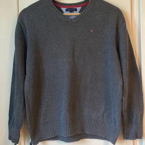 Grå Tommy Hilfiger tröja - Grå stickad tröja från Tommy Hilfiger med v-ringning och diskret logga på bröstet. Tröjan har ribbade muddar vid ärmslut och nederkant samt en snygg röd detalj på insidan av kragen. Storlek 12 men funkar för ålder : 11-14
