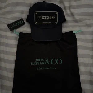 Svart keps Consigliere John Hatter & Co - Svart keps från John Hatter & Co med metallskylt framtill där det står 'CONSIGLIERE'. Klassisk trucker-modell med mesh på sidorna och justerbar baksida. Snygg och stilren accessoar som sticker ut med sin tydliga skylt.