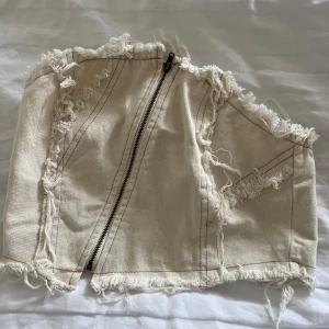 Beige jeanstopp med fransar - Säljer en beige korsettopp i jeansmaterial med råa franskantsdetaljer och synliga sömmar. Toppen är croppad och har dragkedja framtill för en edgy look. Perfekt för dig som vill sticka ut med en cool och trendig vibe.🤍 Aldrig använd & skickas inom 1-2 dagar‼️