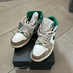 Nike Air Jordan 1 Mid vit/grön mocka - Nike Air Jordan 1 Mid med vit läderbas, grå mockadetaljer och gröna accenter på swoosh, häl och foder. Klassisk snörning och perforerad tå för extra ventilation. Snygg retrostil som sticker ut med sin färgkombination. Använda ganska mycket men eftersom att jag använt crease skydd så ser dom fortfarande bra ut, köpta från merch sweden.