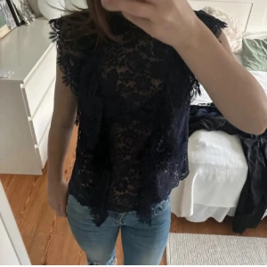 Mörkblå spetsblus med volang - Supersnygg mörkblå blus i genombruten spets med blommigt mönster och volangdetaljer framtill. Ärmlös modell med hög hals och fina kanter. Perfekt att styla med jeans eller kjol för en trendig look.