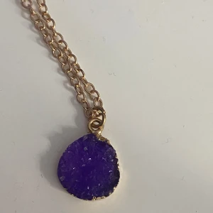 Halsband med lila kristallhänge - Unikt halsband med en chunky kedja i guldton och ett rått, lila kristallhänge. Hänget har en oregelbunden form och glittrar snyggt i ljuset. Perfekt statement-piece för dig som vill sticka ut med färg och coola detaljer.