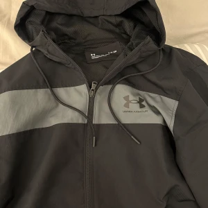 Svart vindjacka från Under Armour - Säljer en stilren svart vindjacka från Under Armour med grå detaljer. Jackan har en dragkedja framtill och en justerbar huva. Perfekt för blåsiga dagar och sportiga aktiviteter.