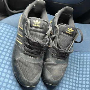 Svarta Adidas Le Trainer sneakers - Säljer ett par svarta Adidas Le Trainer sneakers med guldiga detaljer och klassiska tre ränder på sidorna. Skorna har snörning, platt sula och är tillverkade i syntetmaterial och mesh för en sportig look. Adidas logga syns både på plös och häl.