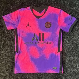 PSG x Jordan rosa-lila fotbollströja - Snygg PSG fotbollströja i samarbete med Jordan. Tröjan har ett unikt rosa och lila mönster med svarta detaljer, klubbmärke på bröstet och Jordan-logga. Tillverkad i lätt och ventilerande material, perfekt för träning och sommaren.