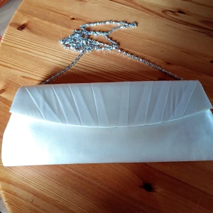 Vit kuvertväska i satin med kedja - Stilren vit kuvertväska i glansigt satinmaterial med elegant veckad framsida och silverfärgad kedja. Perfekt att bära över axeln eller i handen för en extra touch till din outfit.