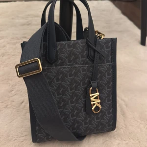 Blå axelväska från Michael Kors - Snygg blå axelväska från Michael Kors med MK-mönster och guldfärgade detaljer. Väskan har både handtag och bred axelrem, samt ett dekorativt MK-hänge framtill. Tillverkad i slitstarkt syntetmaterial med stilren, fyrkantig form. Aldrig använt! 