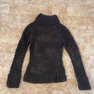 Mörkbrun stickad polotröja - Säljer en mörkgrå stickad polotröja med grovt virkat nätmönster. Tröjan har hög polokrage, långa ärmar och breda ribbade muddar vid ärmslut och nederkant. 