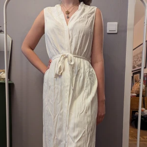 Vit ärmlös långklänning med knyt - Snygg vit långklänning med v-ringning och knytband i midjan. Klänningen är ärmlös och har en avslappnad passform, perfekt för varma dagar.