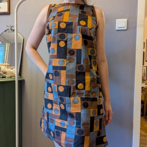 Retro mönstrad ärmlös klänning - Säljer en handgjord ärmlös klänning med retroinspirerat geometriskt mönster i brunt, gult, svart och blått. Klänningen är kort och har en rak passform med rund halsringning. 