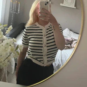 Randig kortärmad kofta  - Supersöt kortärmad kofta i vitt och svart med horisontella ränder. Tröjan har pärlknappar framtill, ribbade kanter och är croppad i modellen. Perfekt för dig som gillar en klassisk men trendig look. Ny med prislapp 