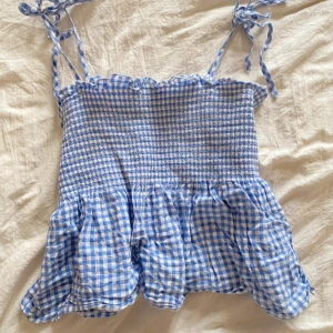 Blåvit rutig linnetopp från H&M - Supersöt blå och vit rutig linnetopp från H&M med smockad överdel, volang nertill och knytbara axelband. Perfekt för varma dagar och ger en riktigt somrig vibe. Tunt och lätt material som känns luftigt och skönt.
