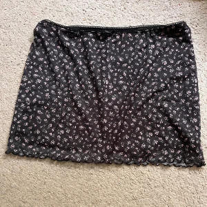 Svart blommig kortkjol med spetskant - Supersöt svart kortkjol med små blommor i ljusrosa och vit. Kjolen har en fin spetskant nertill och en smal spetsdetalj upptill i midjan. Funkar som top också! Aldrig använd