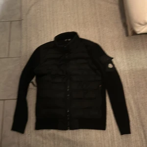 Svart Moncler pufferjacka med dragkedja - Snygg svart pufferjacka från Moncler med quiltade partier framtill och på axlarna. Jackan har lång ärm, hög krage och dragkedja hela vägen. Ikonisk Moncler-logga på ena ärmen. Perfekt för dig som vill ha en stilren och exklusiv look.
