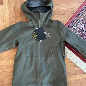 Arc’teryx Beta LT - Säljer en stilren mörkgrön skaljacka modell Beta LT från Arc'teryx i GORE-TEX-material. Jackan har en justerbar huva och flera praktiska dragkedjefickor. Perfekt för regniga dagar och utomhusaktiviteter som vandring eller skidåkning.