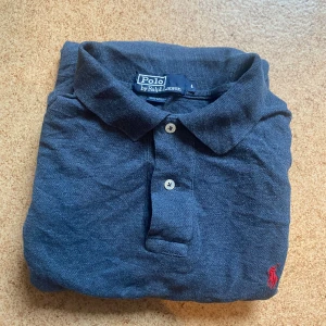 Mörkblå långärmad piké från Ralph Lauren - Snygg mörkblå långärmad pikétröja från Polo Ralph Lauren med klassisk krage, två knappar och röd broderad logga på bröstet. Tillverkad i mjuk bomull, perfekt för en chill och stilren look.