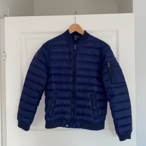 Marinblå dunjacka Polo Ralph Lauren - Snygg marinblå dunjacka från Polo Ralph Lauren med klassisk bombermodell. Dragkedja framtill, fickor med tryckknappar och en ärmficka med zip. Tydlig logga på ärmen. Litet hål på ryggen men lätt att sy ihop.
