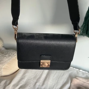 Svart axelväska med gulddetalj - Stilren svart axelväska i skinnimitation med bred axelrem och snyggt guldfärgat spänne framtill. Perfekt storlek för mobil, plånbok och andra essentials. Enkel och clean design som passar till många outfits.